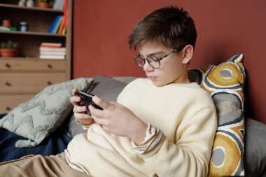 Gözlüklü ve kazaklı bir öğrenci kameranın önünde oturmuş bilgisayar oyunu oynarken akıllı telefon ekranına bakıyor.