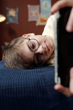 Gözlüklü sevimli sarışın çocuk ev ortamında yatakta uzanıyor ve akıllı telefonuyla oyun oynuyor.