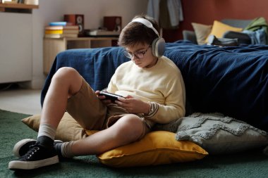 Günümüz okul çocuğu yatağın yanında oturmuş oyun alanındaki cep telefonu ekranına bakarken video oyunu oynuyor.