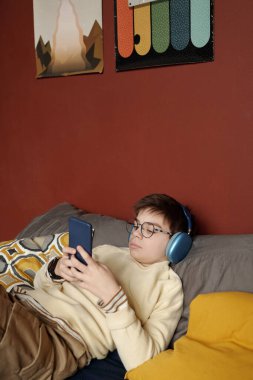 Ciddi okul çocuğu, gündelik giysiler ve kulaklıklar takıyor. Yatakta dinlenip video izlerken elinde akıllı telefon ekranına bakıyor.