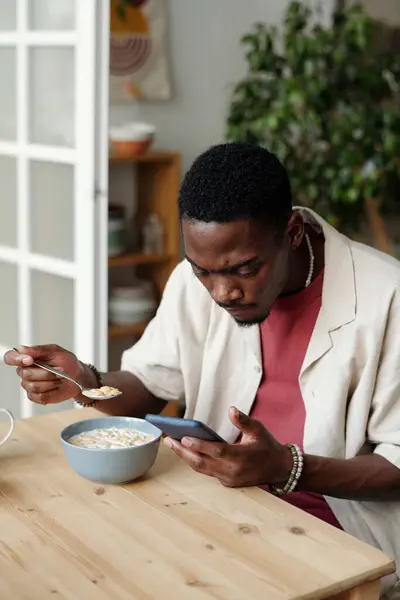 非洲裔美国人坐在餐桌旁 一边吃着牛奶玉米片 一边看在线视频 一边用手机发短信 — 图库照片