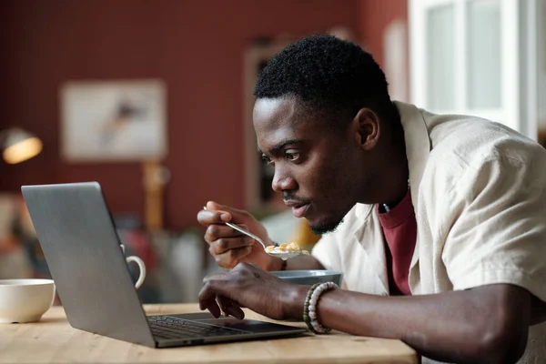 Young man looking at laptop screen while watching online video or communicating with someone in chat during breakfast