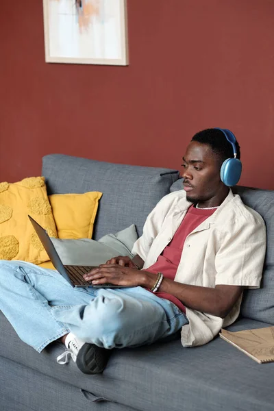 カジュアルウェアとヘッドフォンで若い現代アフリカ系アメリカ人のビジネスマンがソファに座ってオンラインデータを分析 — ストック写真