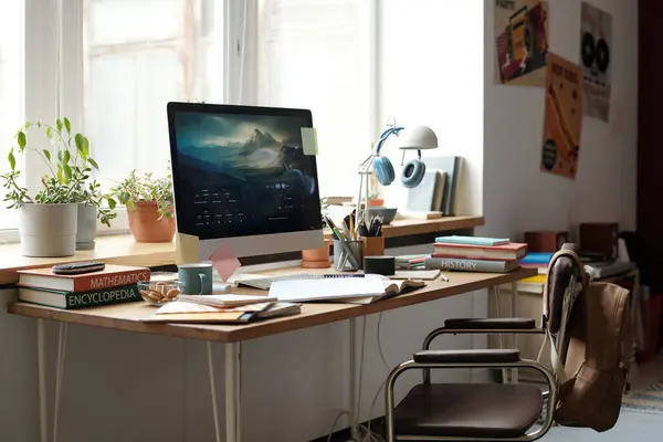 Workplace of solopreneur, student or designer with computer monitor, books, notepads ang group of flowerpots standing along windowsill
