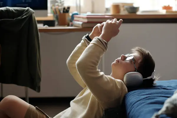 ベッドに座って ビデオゲーム中にゲームパッドに接続されたモバイルガジェットの画面を見ているヘッドフォンの若い男の子のサイドビュー — ストック写真