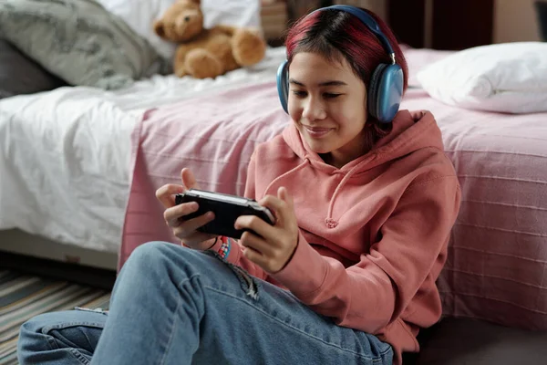 幸せな十代の少女とゲームパッドのスマートフォンは 彼女のベッドで床に座って ビデオゲームをプレイしながら画面を見ています — ストック写真