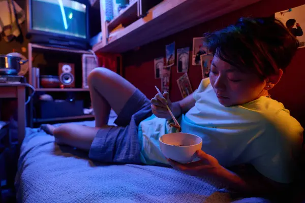 カメラの前でシングルベッドでリラックスしたアジアの少女が小さなアパートでボウルから中国料理を食べる — ストック写真