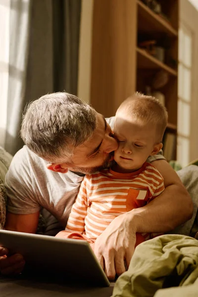 Affectionate father kissing his adorable baby son on cheek and embracing him while both relaxing at leisure and watching online video
