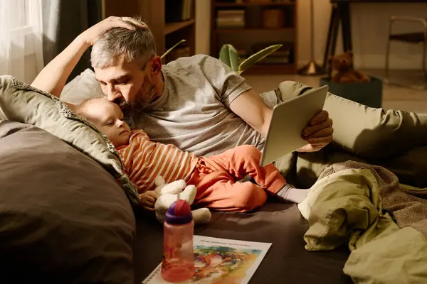 彼の隣に横たわる彼の愛らしい赤ちゃんの息子にキスし 成熟した父親が保持するタブレットの画面を見ている灰色の髪を持つ男 — ストック写真