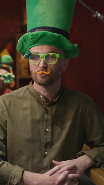 セントパトリックのお祝いの日の間にアイルランドのバーでカメラのための偽の口ひげと面白い眼鏡を身に着けている白人男性の垂直の肖像画 — ストック動画