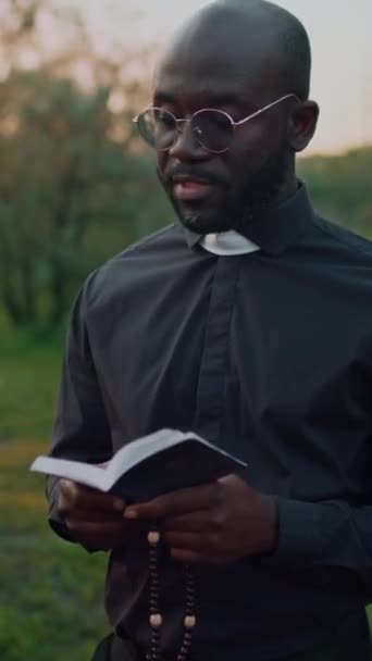 アフリカ系アメリカ人の司祭のメガネの中央垂直映像は 埋葬サービスの読書の祈りの本と屋外の葬儀でビーズを保持しています — ストック動画