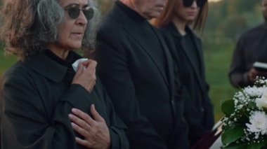 Güneş gözlüklü yaşlı beyaz bir kadının yan görüntüsü. Ağlıyor ve mendil kullanıyor. Açık hava cenaze töreninde tahta tabutun yanında akrabalarının yanında duruyor.
