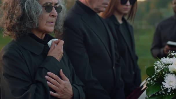 サングラスの上級白人女性が涙を流し 屋外の葬儀で木製の棺で親戚と一緒に立っているハンカチを使用して傾いた側面映像 — ストック動画