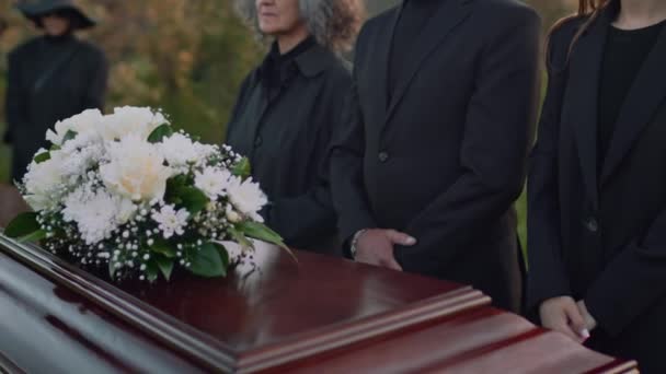 黒い服とサングラスの親戚のチルト側の映像は 屋外での葬儀サービス中に蓋に白い花が付いた木製のケケットの近くに立っています — ストック動画