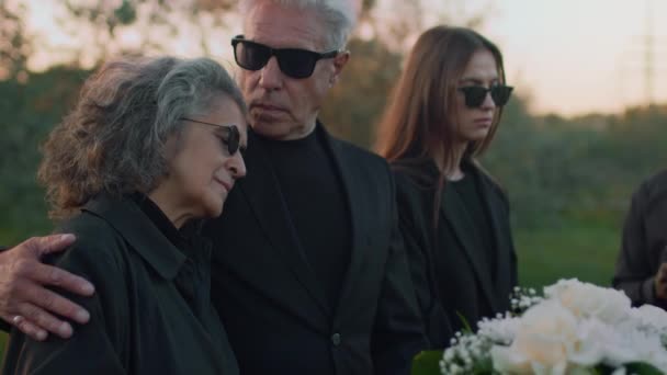 死者を悼む涙を抱く白人上級女性の側面のメディア映像と屋外の葬儀で木棺で彼女の立場を慰める男 — ストック動画