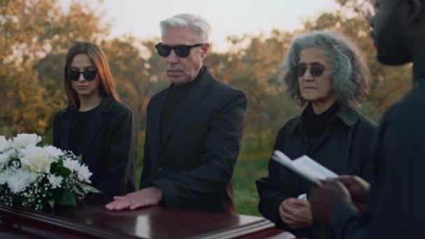 黒い司祭の葬儀の間に墓地の親戚によって立っているそれの上に白い花が付いている木製のカスケットのふたに手を置く上級白人男性の側面媒体の映像 — ストック動画