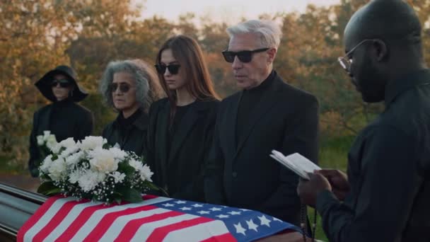 アフリカ系アメリカ人の司祭が葬儀サービスを読んでおり 高齢の白人男性が墓地に家族と一緒に立っている間に亡くなったとさよならを言って 米国国旗を掲げた — ストック動画