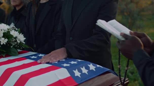 サングラスのコーカサス上級男性のチルト側の映像は 墓地での記念礼拝中に米国の埋葬旗で覆われた木製棺の蓋に手を置きます — ストック動画