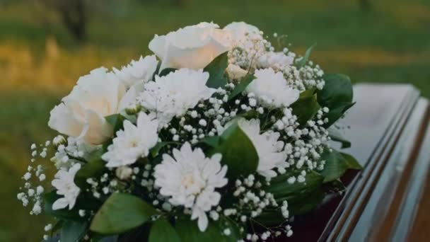 墓地での葬儀中に木製の棺の蓋に横たわる白いバラと菊の花束と認識できない手のチルト側のクローズアップ — ストック動画