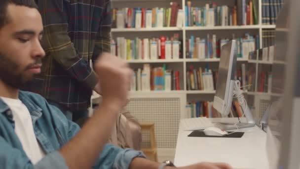大学図書館のデスクトップコンピュータに座っている忙しいアラブの男性学生のミディアムクローズアップショット キーボードの思考とタイピング 認識できない男性が入ってくる ノートブックを取り出して作業する準備 — ストック動画