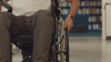 Tekerlekli sandalyedeki engelli genç Orta Doğulu erkek öğrencinin, kütüphane sıraları arasında gezinip, çalışmak için ders kitapları ararken orta eğik çekimleri.