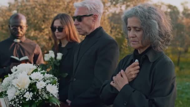 墓地で黒人の司祭が行った記念式典の間 木製のケケットで家族と一緒に祈り泣いている白い女性の側面の中間映像 — ストック動画