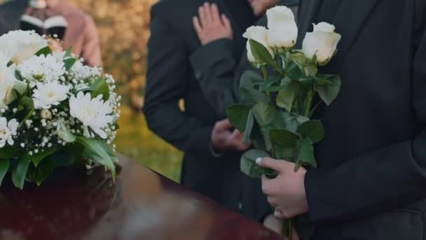 木の棺に立って 屋外の葬儀で白いバラを手に持っている 認識できない女性の側面映像 — ストック動画