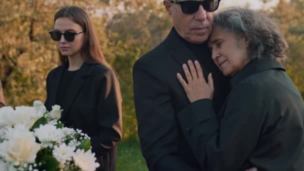 墓地で彼女の息子や娘の葬儀で悲しんでいる女性を慰めるコーカサス上級夫のティルト映像 — ストック動画