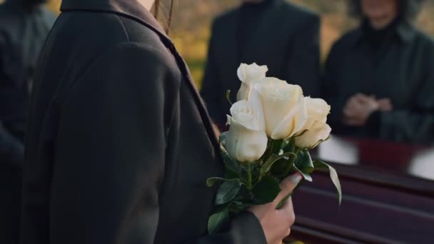 家族の近くの木製棺で白いバラと立っている若い白い白いバラの若い白い女性のキルト側のクローズアップと墓地で悲しみを感じる — ストック動画