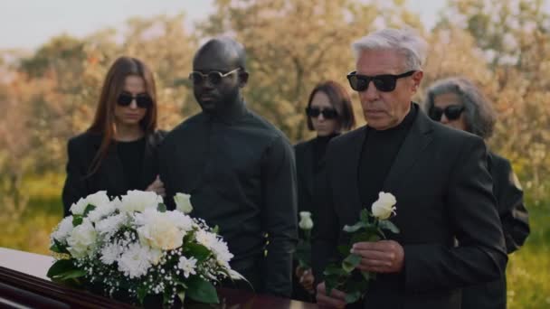 白いバラの花が1本の白いバラと一緒に立っているコーカサス人のシニアの側面の映像は 墓地の多様な家族と別れの葬儀で 木製のカスケットで — ストック動画