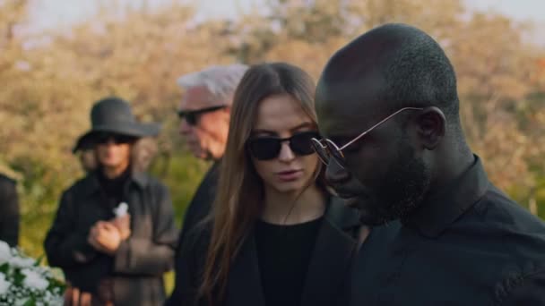 アフリカ系アメリカ人の男性を泣かせるためにサングラスにいた白人女性のサイドチェスト 屋外の葬儀で哀悼の意を表明 — ストック動画