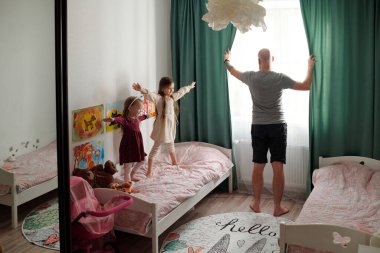 İç çamaşırlı genç kel bir adamın sabah yeşil perdeleri açarken iki kızının yanında spor yaparken görüntüsü.