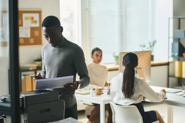 若いアフリカ系アメリカ人のオフィスマネージャーは 同僚に対するXeroxマシンによって立っている間 財務文書を見ています — ストック写真