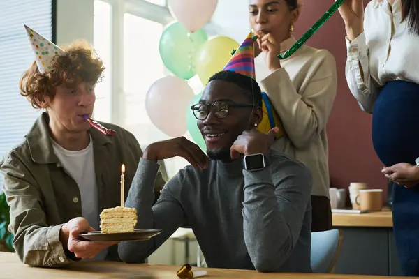 Happy young man in birthday cap looking at piece of tasty cake with burning candle while sitting by table among colleagues