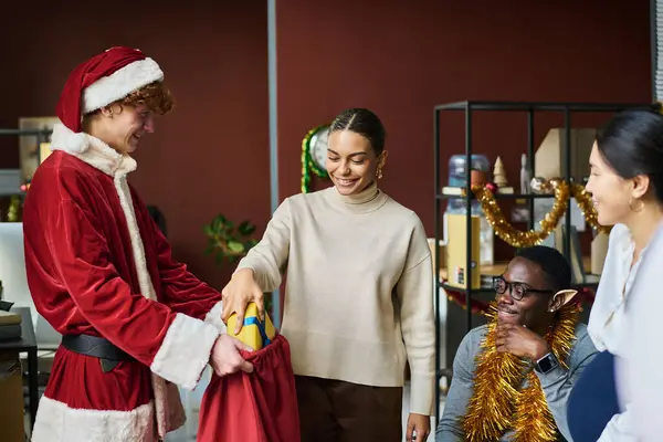 若い笑顔のアフリカ系アメリカ人ビジネスマンは サンタクロースの衣装を着た男性同僚が保持する赤い袋からギフトボックスを取り出します — ストック写真