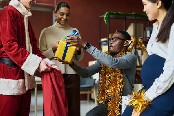 幸せな若いアフリカ系アメリカ人男性は サンタのコスチュームで男が保持するXmasプレゼントで赤い袋から引き出されたギフトボックスを保持しています — ストック写真