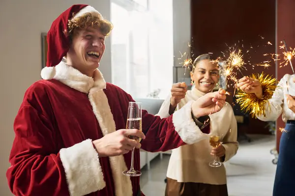 サンタクロースのコスチュームで若い笑うビジネスマンは Xmasのお祝いを楽しんでいる間 火花とシャンパンのフルートを保持しています — ストック写真