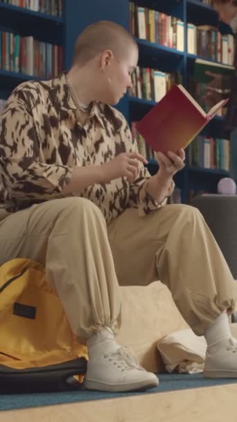 カジュアルな服を着た白人女性学生の完全な縦のショット 大学図書館に座って剃られた頭部 データ分析の教科書を読み そしてスマートフォンで講演する時間を見る — ストック動画
