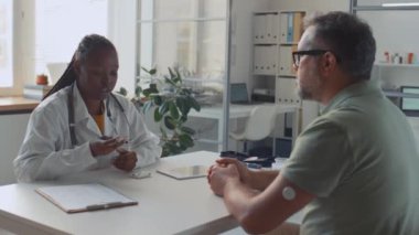 Orta yaşlı beyaz bir hastaya danışmak ve kan şekeri kontrol metodu göstermek için klinikte masa başında oturan Afrikalı Amerikalı kadın doktorların orta boy fotoğrafları.