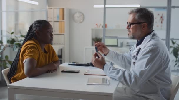 診療所で糖尿病を患う新しいアフリカ系アメリカ人女性患者に相談する男性医師の中型ショット 血糖コントロールのためのインスリンペンを示し 薬を注入する方法を教える 女性のリスニング — ストック動画