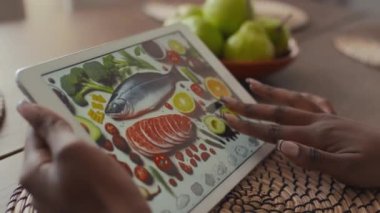 Afro-Amerikalı bir kadının elleri tablet bilgisayarında diyabetik gıdalar, taze sebzeler, balıklar, et, avokado, brokoli ile fotoğrafa bakıyor, büyütüyor ve dokunmatik ekranda kaydırıyor.