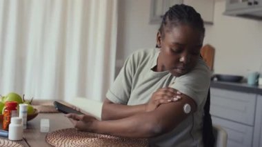 Orta boy Afro-Amerikan kadın diyabetli evde mutfak masasında oturuyor, kolundaki glikoz izleme cihazına bakıyor, sonra akıllı telefon uygulamasındaki verileri kontrol ediyor ve memnuniyetle başını sallıyor.