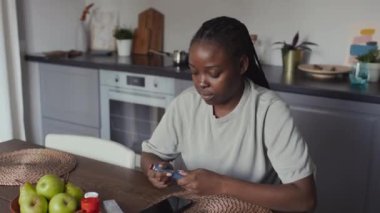 Orta ölçekli örülü saçlı, günlük tişörtlü, mutfakta oturan, insülin kalemini açan ve koluna iğne yapan, ilaç kutuları ve meyveleri masaya koyan Afro-Amerikan kadının fotoğrafı.