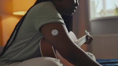 Orta ölçekli diyabet hastası genç siyahi bir kadının koluna kan şekeri monitörü takıp, yatak odasında otururken, dizüstü bilgisayarını açarken, gitar çalarken ve şarkı söylerken.