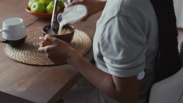 キッチンテーブルに座って腕にブドウ糖モニター装置を装備した認識できないアフリカ系アメリカ人女性のミディアムリアショット 健康的な低血糖朝食のためのドライオートフレークのボウルにミルクを注ぐ — ストック動画