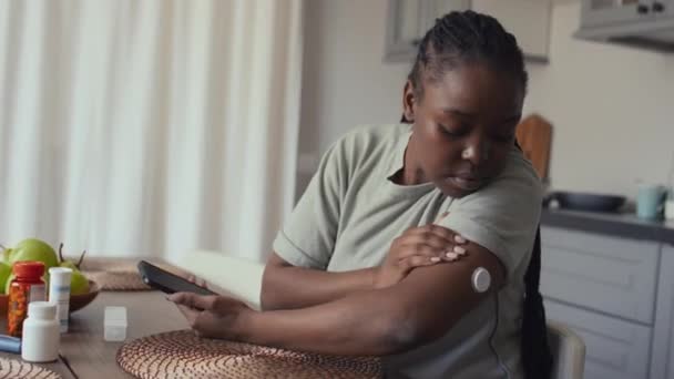 自宅でキッチンテーブルに座って糖尿病を患っているアフリカ系アメリカ人女性の中型ショット 腕のグルコースモニタリングデバイスを見て スマートフォンアプリのデータをチェックし 満足してうなずく — ストック動画