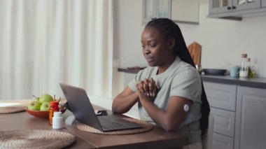 Orta boy gülümseyen, şeker hastası bir Afro-Amerikan kadını dizüstü bilgisayarın önünde mutfak masasında otururken, arkadaşıyla video görüşmesi yaparken, sohbet ederken ve kolunda yeni glikoz izleme cihazını gösterirken.