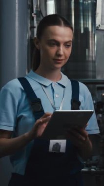 Polo gömlekli ve tulumlu genç beyaz kadın mühendisin su şişeleme fabrikasında tablet kullanarak şişe üretim sürecini incelediği dikey bıçak görüntüleri.