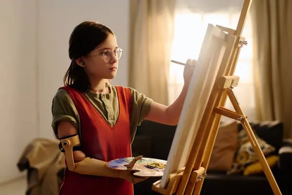 Diligent schoolgirl with hand prosthesis standing in front of easel with unfinished painting in living room and creating new artwork