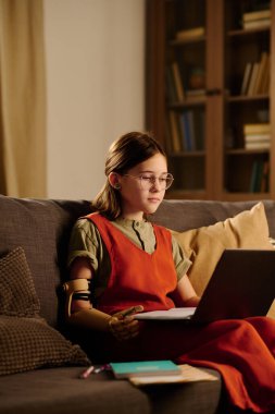 Çalışkan ampüte genç kız oturma odasında kanepede oturuyor ve online ders sırasında dizlerindeki dizüstü bilgisayara bakıyor.